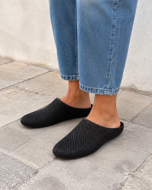 A person wearing black mesh slip-on mule shoes with blue jeans, standing on a gray stone pavement against a light-colored wall.