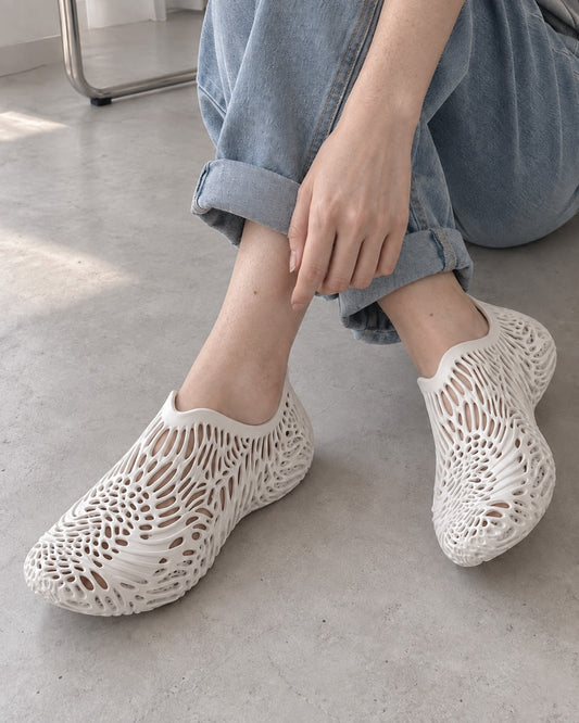 A person wearing off-white, openwork mesh slip-on shoes with a textured, geometric pattern, paired with light blue jeans on a concrete floor.