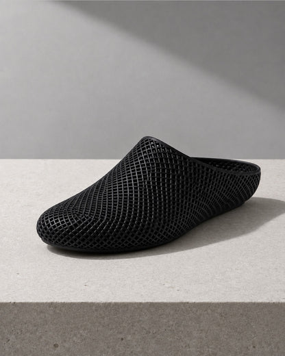 A black mesh slip-on clog shoe with a pointed toe, displayed on a light gray concrete surface against a gray background with a diagonal shadow.