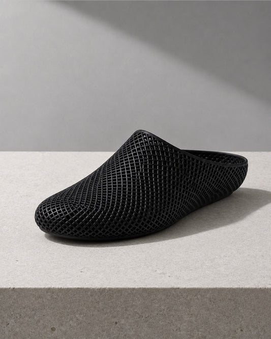 A black mesh slip-on clog shoe with a pointed toe, displayed on a light gray concrete surface against a gray background with a diagonal shadow.