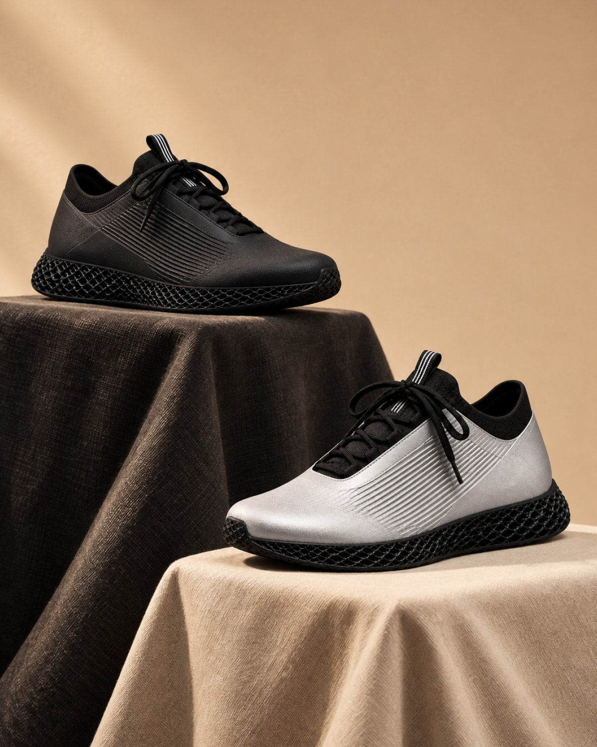 Two low-top athletic sneakers displayed on draped fabric pedestals against a warm beige studio background. The upper shoe is black and sits on a dark brown cloth, while the lower shoe is white with black detailing and sits on a beige cloth.