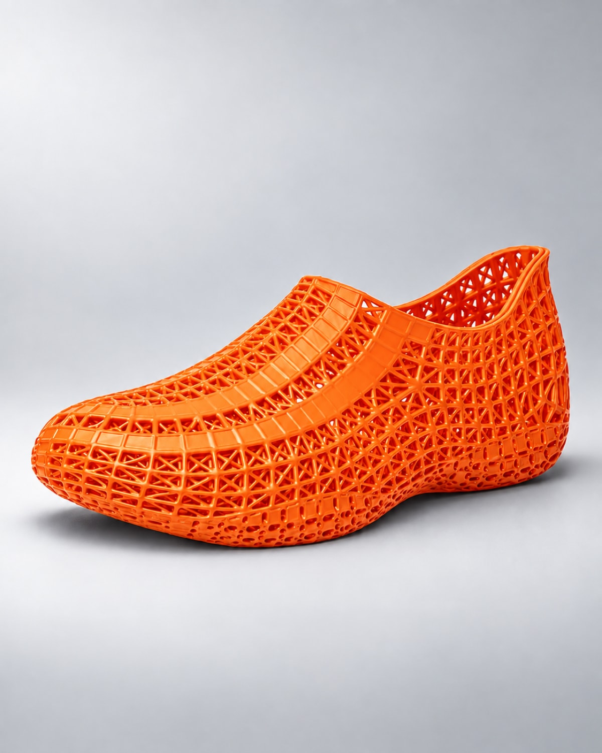 A bright orange 3D-printed slip-on shoe with an intricate open-weave geometric pattern, shown from a side profile on a light gray background.