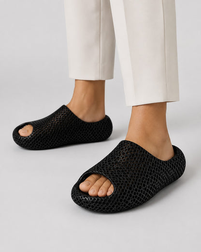 A person wearing black open-toe mesh slip-on shoes with a textured, reptile-like pattern, paired with off-white trousers, shown against a light gray studio background.
