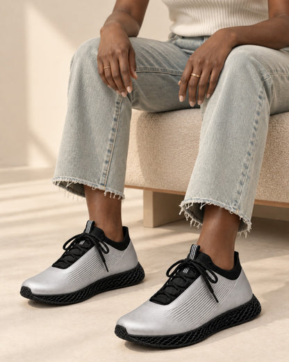 A person seated on a beige bench wearing metallic silver and black athletic sneakers with light-wash frayed-hem jeans in a softly lit indoor setting.