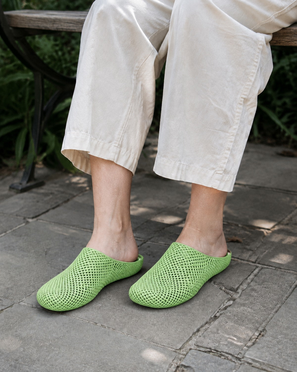 A person sitting on a bench outdoors wearing light beige pants and bright green open-toe mule slippers on a stone patio.