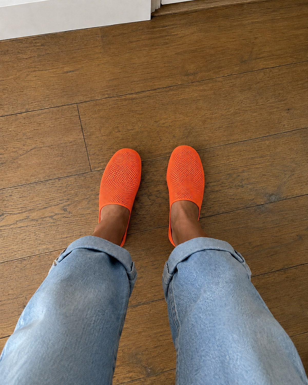 A person wearing bright orange slip-on mesh shoes with cuffed light blue jeans, standing on a brown wooden floor.