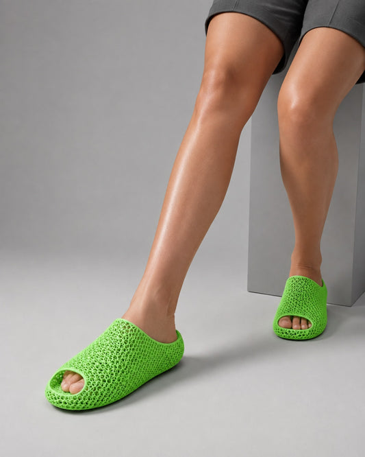 A person wearing bright neon green open-toe crochet-style slippers, shown from the thighs down against a light gray studio background with a gray pedestal.