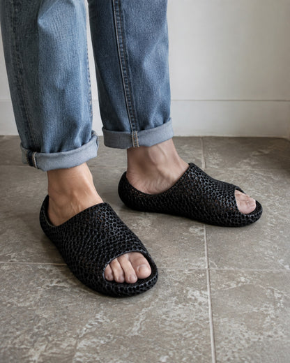 Black woven open-toe slide sandals worn with cuffed blue jeans on a tiled floor.