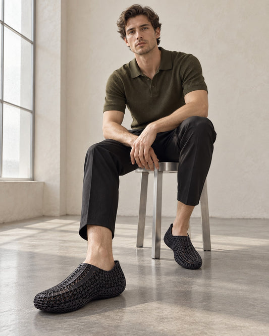 A man sits on a metal stool in a bright, modern room, wearing a dark olive green polo shirt, black trousers, and black woven slip-on shoes.