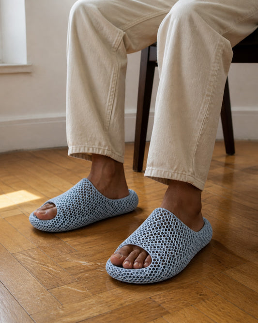 A person seated indoors wearing light blue open-toe crochet slippers with cream pants, standing on a wooden floor.