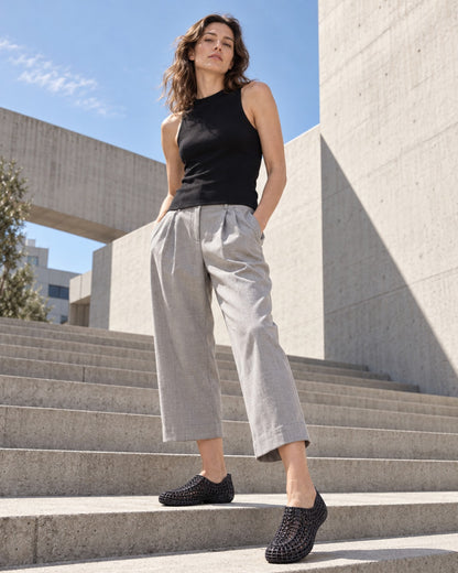 A woman stands on outdoor concrete stairs wearing a black sleeveless top, light gray cropped wide-leg pants, and black woven slip-on shoes.