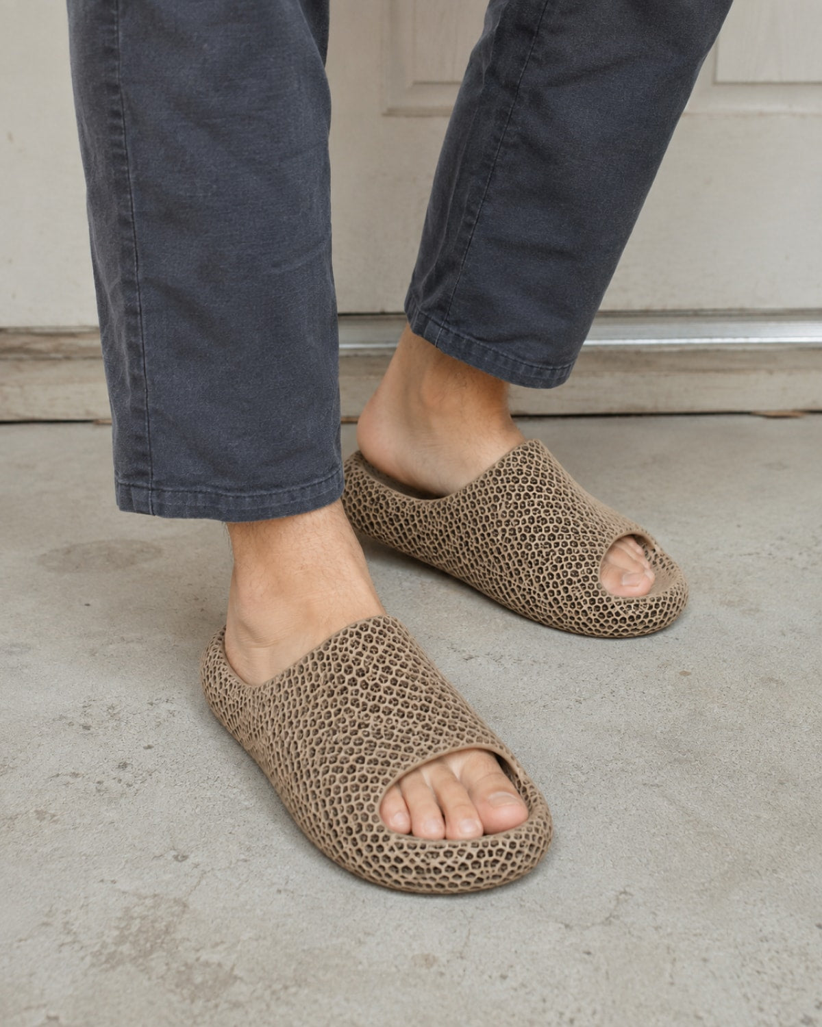 A person wearing brown open-toe crochet knit slip-on slippers with dark gray jeans, standing on a concrete floor.