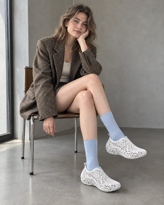 A woman sits on a wooden chair in a minimalist room, wearing a brown tweed blazer, white top, light blue socks, and white slip-on shoes with a geometric knit pattern.