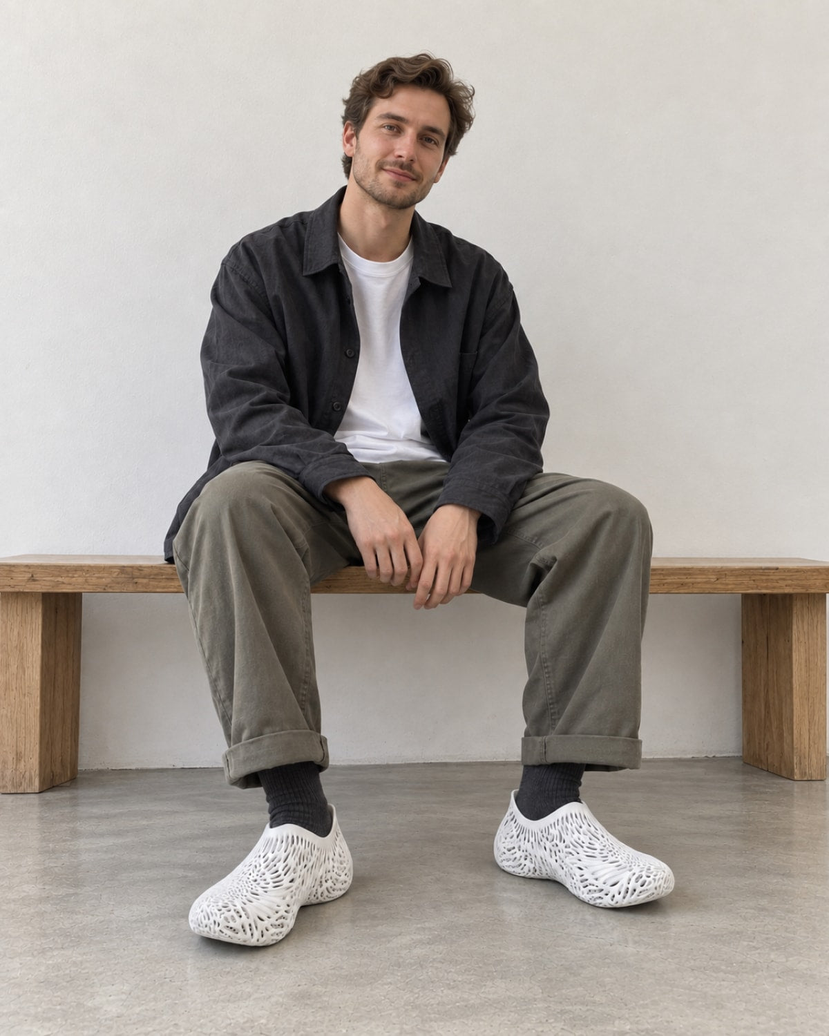 A man sits on a wooden bench in a minimalist room, wearing a dark charcoal overshirt, olive green trousers, black socks, and distinctive white openwork mesh slip-on shoes.