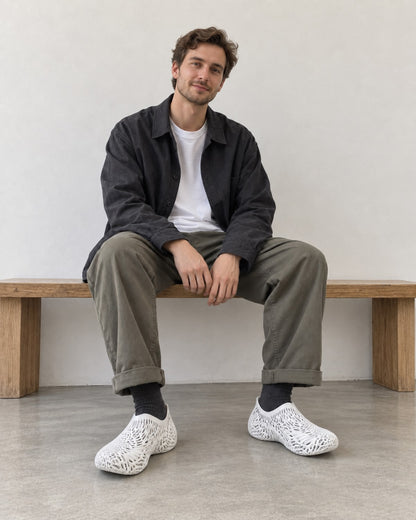 A man sits on a wooden bench in a minimalist room, wearing a dark charcoal overshirt, olive green trousers, black socks, and distinctive white openwork mesh slip-on shoes.